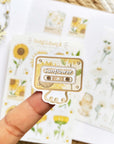 Premium Handmade Sticker Sheet Chamomile Sunflower 105mm x 148mm - a piece on the finger and 2 sheets on the table with a white flower branch, an opened notebook