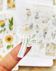 Premium Handmade Sticker Sheet Chamomile Sunflower 105mm x 148mm - a piece on the finger and 2 sheets on the table with an opened notebook