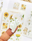 Premium Handmade Sticker Sheet Chamomile Sunflower 105mm x 148mm - a piece on the finger and 2 sheets on the table with a white flower branch, an opened notebook