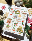 Premium Handmade Sticker Sheet Christmas 105mm x 148mm - a sheet on the table with a white flower branch, an opened notebook, 3 pinecones, 2 pine branches, 2 red berry branches, 5 stars
