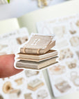 Premium Handmade Sticker Sheet Dark & Light Academia 105mm x 148mm - a piece on the finger and 2 sheets on the table with an opened notebook