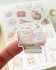 Premium Handmade Sticker Sheet Knitting Crochet 105mm x 148mm - a piece on the finger and a sheet on the table with an opened notebook