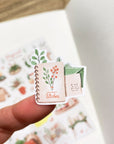 Premium Handmade Sticker Sheet Magical Cozy Winter 105mm x 148mm - a piece and a sheet on the table with an opened notebook