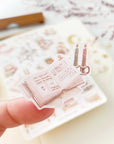 Premium Handmade Sticker Sheet Reading at Home 105mm x 148mm - a piece on the finger and a sheet on the table with a dark maroon flower branch, an opened notebook