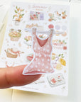Premium Handmade Sticker Sheet Summer Breeze 105mm x 148mm - a piece on the finger and a sheet on the table with an opened notebook