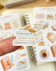 Premium Handmade Sticker Sheet Aesthetic Book 105mm x 148mm - a piece on the finger and 2 sheets on the table with an opened notebook