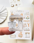 Premium Handmade Sticker Sheet Aesthetic Cozy Kitchen 105mm x 148mm - a piece on the finger and a sheet on the table with a pencil, an opened notebook