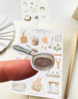 Premium Handmade Sticker Sheet Aesthetic Cozy Kitchen 105mm x 148mm - a piece on the finger and a sheet on the table with a pencil, an opened notebook
