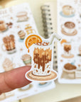 Premium Handmade Sticker Sheet Coffee 105mm x 148mm - a piece on the finger and a sheet on the table with an opened notebook