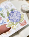 Premium Handmade Sticker Sheet Floral Power 105mm x 148mm - a piece on the finger and a sheet on the table with a white flower branch, an opened notebook, a washi tape
