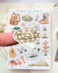 Premium Handmade Sticker Sheet Into The Wild 105mm x 148mm - a piece on the finger and a sheet on the table with a white flower branch, an opened notebook, a washi tape