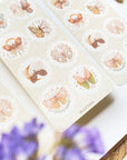 Premium Handmade Sticker Sheet Magic Wings 105mm x 148mm - 2 sheets on the table with a purple flower branch, an opened notebook