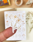 Premium Handmade Sticker Sheet Mellow Flower 105mm x 148mm - a piece on the finger and a sheet on the table with a white flower branch, an opened notebook