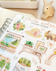 Premium Handmade Sticker Sheet Spring Garden 105mm x 148mm - a sheet on the table with a rabbit statue, an opened notebook