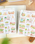 Premium Handmade Sticker Sheet Spring Garden 105mm x 148mm - 2 sheets on the table with a rabbit statue, an opened notebook