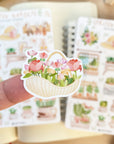 Premium Handmade Sticker Sheet Spring Garden 105mm x 148mm - a piece on the finger and 2 sheets on the table with an opened notebook