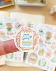 Premium Handmade Sticker Sheet Spring Vibes 105mm x 148mm - a piece on the finger and 2 sheets on the table with an opened notebook