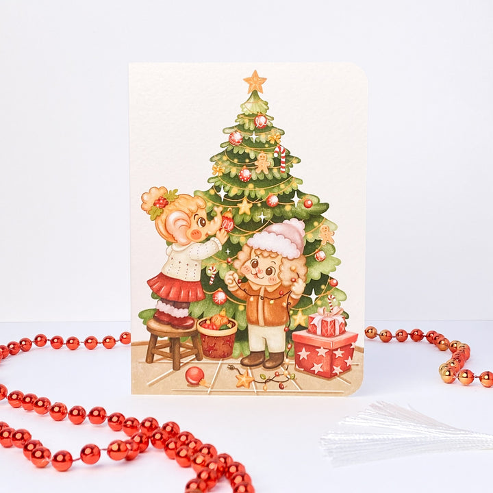 Decorating Tree Christmas card without text: Tuk Tuk the elephant hangs baubles while Stroop the dog holds the lights