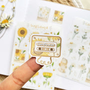 Premium Handmade Sticker Sheet Chamomile Sunflower 105mm x 148mm - a piece on the finger and 2 sheets on the table with a white flower branch, an opened notebook