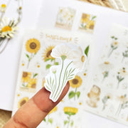 Premium Handmade Sticker Sheet Chamomile Sunflower 105mm x 148mm - a piece on the finger and 2 sheets on the table with a white flower branch, an opened notebook