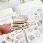 Premium Handmade Sticker Sheet Dark & Light Academia 105mm x 148mm - a piece on the finger and 2 sheets on the table with an opened notebook