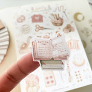 Premium Handmade Sticker Sheet Knitting Crochet 105mm x 148mm - a piece on the finger and a sheet on the table with an opened notebook
