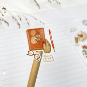 Slow Winter kiss-cut sticker on a pencil: a hand with a red pen writing in a notebook