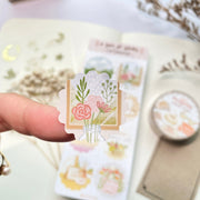Premium Handmade Sticker Sheet A Day At Home 105mm x 148mm - a piece on the finger and a sheet on the table with a white flower branch, a purple flower branch, a washi tape, an opened notebook