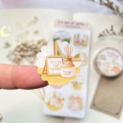 Premium Handmade Sticker Sheet A Day At Home 105mm x 148mm - a piece on the finger and a sheet on the table with a white flower branch, a purple flower branch, a washi tape, an opened notebook