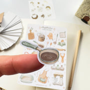 Premium Handmade Sticker Sheet Aesthetic Cozy Kitchen 105mm x 148mm - a piece on the finger and a sheet on the table with a pencil, an opened notebook