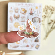 Premium Handmade Sticker Sheet Dark Magic 105mm x 148mm - a piece on the finger and a sheet on the table with a white flower branch, an opened notebook