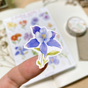 Premium Handmade Sticker Sheet Floral Desire 105mm x 148mm - a piece on the finger and a sheet on the table with a white flower branch, an opened notebook, a washi tape