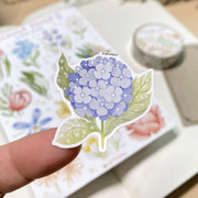 Premium Handmade Sticker Sheet Floral Power 105mm x 148mm - a piece on the finger and a sheet on the table with a white flower branch, an opened notebook, a washi tape