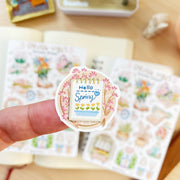 Premium Handmade Sticker Sheet Spring Vibes 105mm x 148mm - a piece on the finger and 2 sheets on the table with an opened notebook